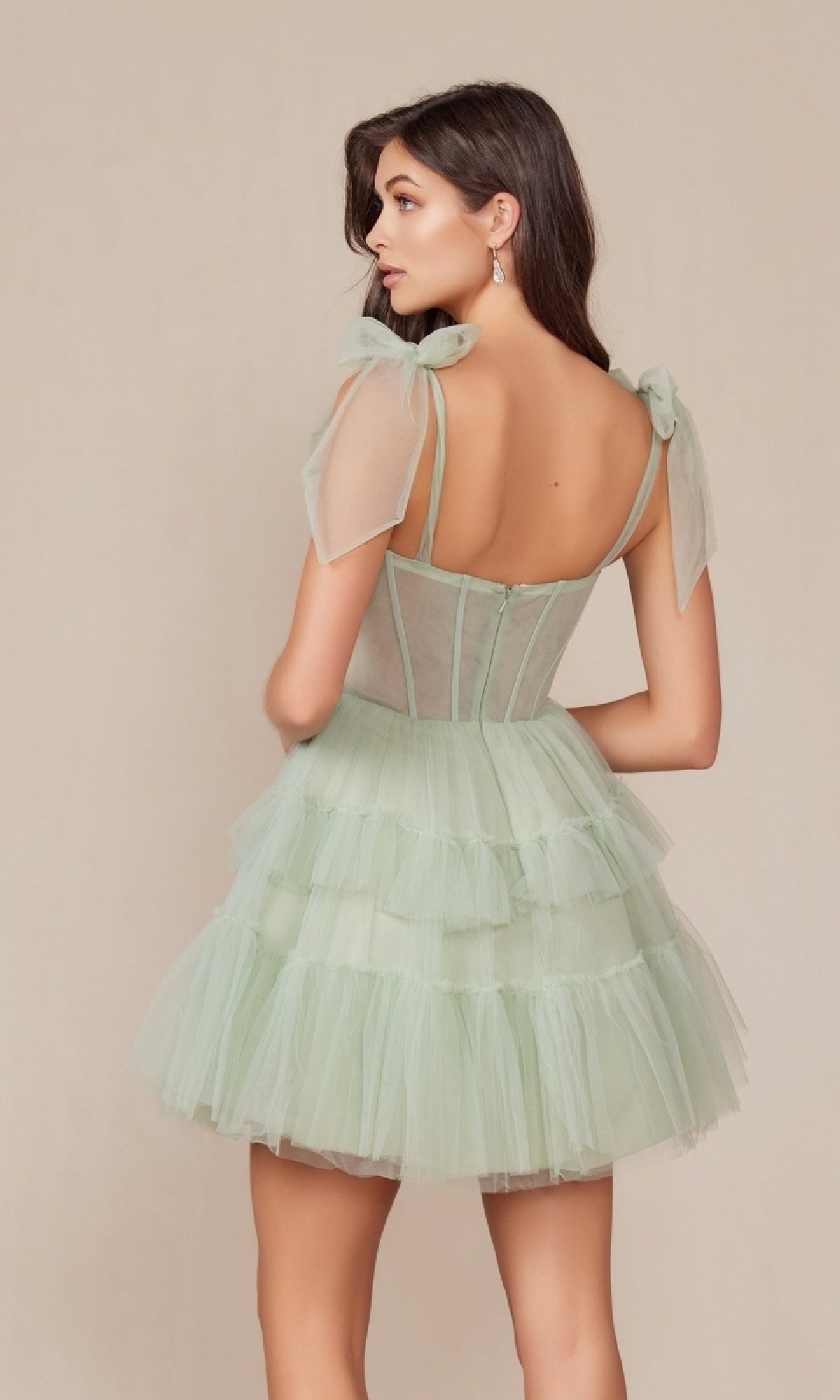 A woman is shown from the back in the Shoulder-Tie Short Ruffled A-Line Hoco Dress Y858, featuring light green sheer bow straps and a tiered tulle skirt, set against a plain beige background.